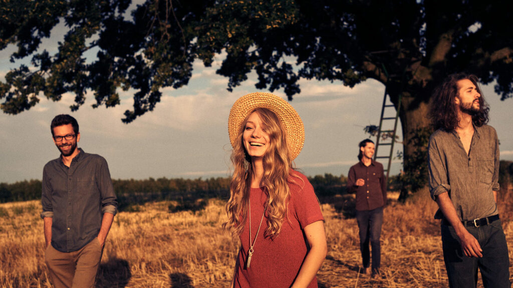 Portraitfoto der Bandmitglieder von Leléka im Feld vor einem Baum, im Vordergrund die Sängerin mit Strohhut.