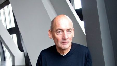 Portraitfoto des niederländischen Architekten Rem Koolhaas, der sich in einem Gebäude an eine Säule lehnt.