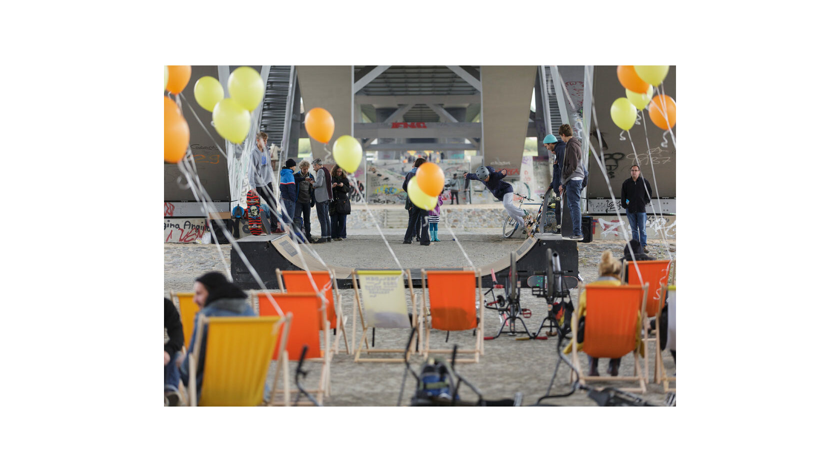 Blick auf eine Minirampe unterhalb der Waldschlösschenbrücke, auf der junge Skater unterwegs sind. Im Vordergrund stehen zahlreiche gelbe und orangene Liegestühle, an denen Luftballons befestigt sind.