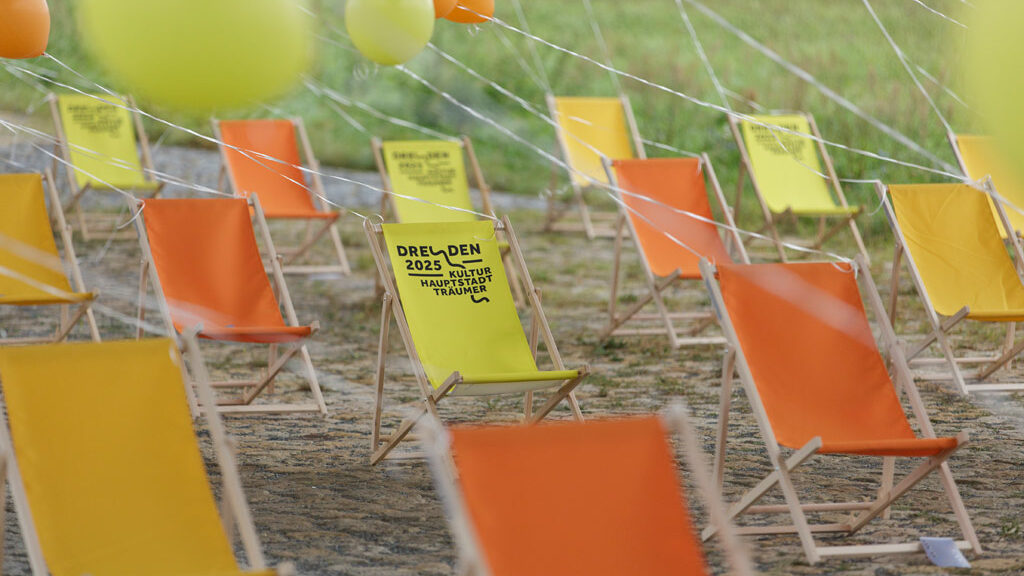 Blick auf zahlreiche gelbe und orangene Liegestühle, an denen gelbe und orangene Luftballons befestigt sind, dazwischen gelbe Liegestühle mit dem Dresden-2025-Logo.