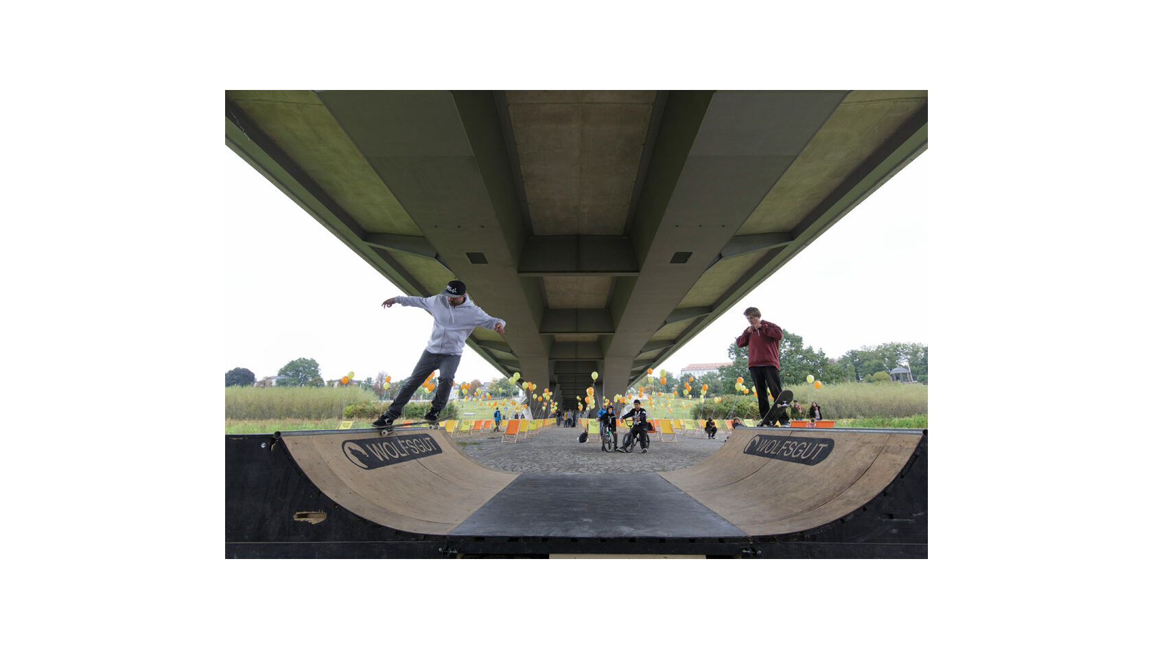 Blick auf eine Minirampe unterhalb der Waldschlösschenbrücke, auf der zwei junge Skater unterwegs sind. Im Hintergrund stehen zahlreiche gelbe und orangene Liegestühle, an denen Luftballons befestigt sind.