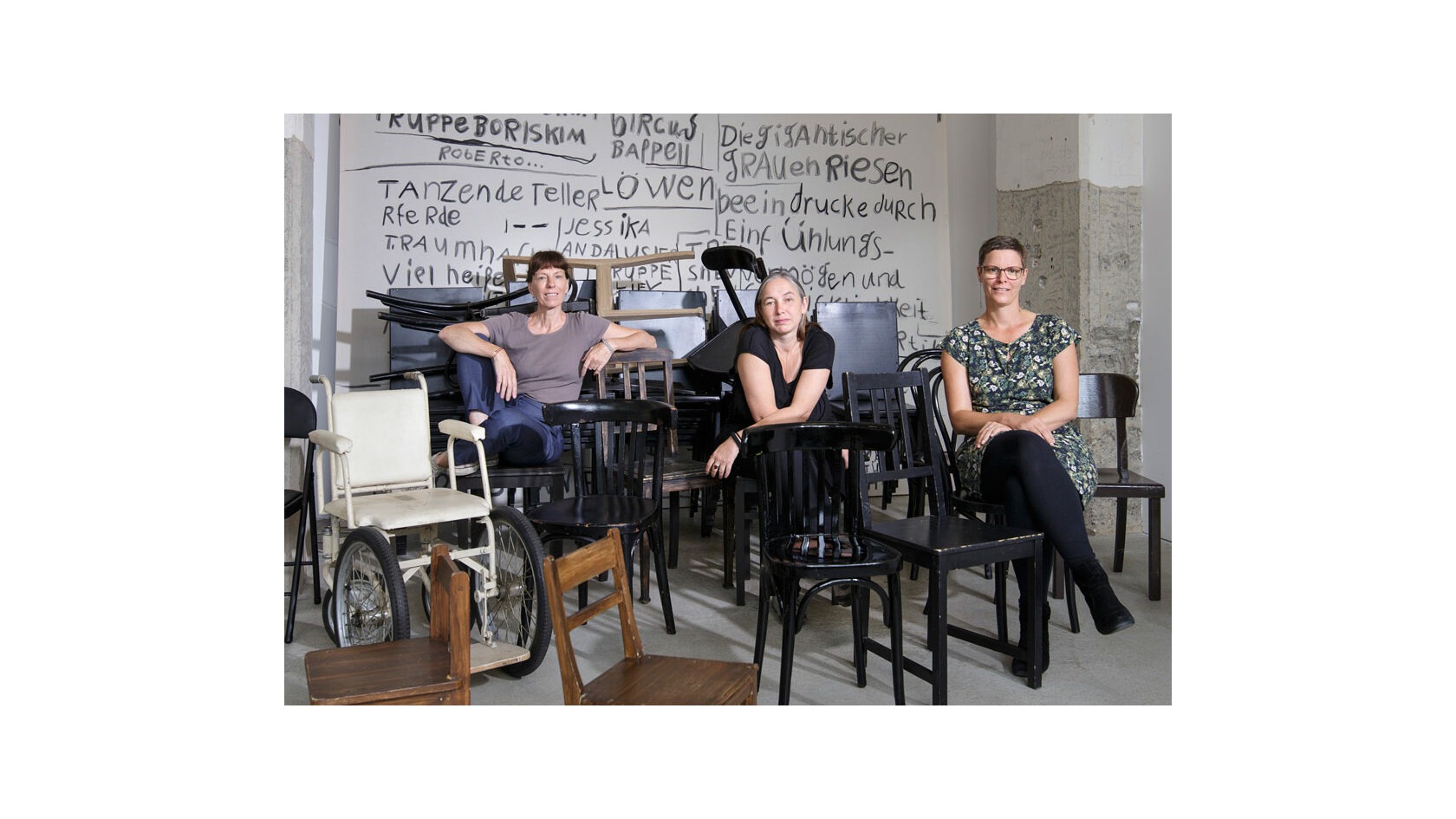 Portraitfoto der Initiatorinnen des "Straßentheaterprojekts" vom Kunst- und Kulturverein farbwerk e. V., Julia Amme, Jacqueline Hamann und Svea Duwe. Sie sitzen in einem Raum voller Stühle neben einem historischen Rollstuhl.