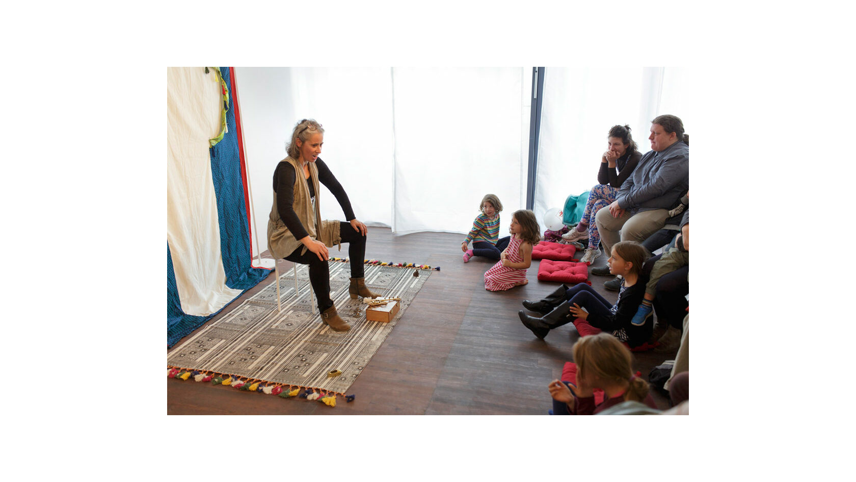 Eine Erzählerin vom Verein Erzählraum e. V. sitzt auf einem ausgerollten Teppich in einem Raum und erzählt vor Kindern und Erwachsenen, die ihr zuhören.