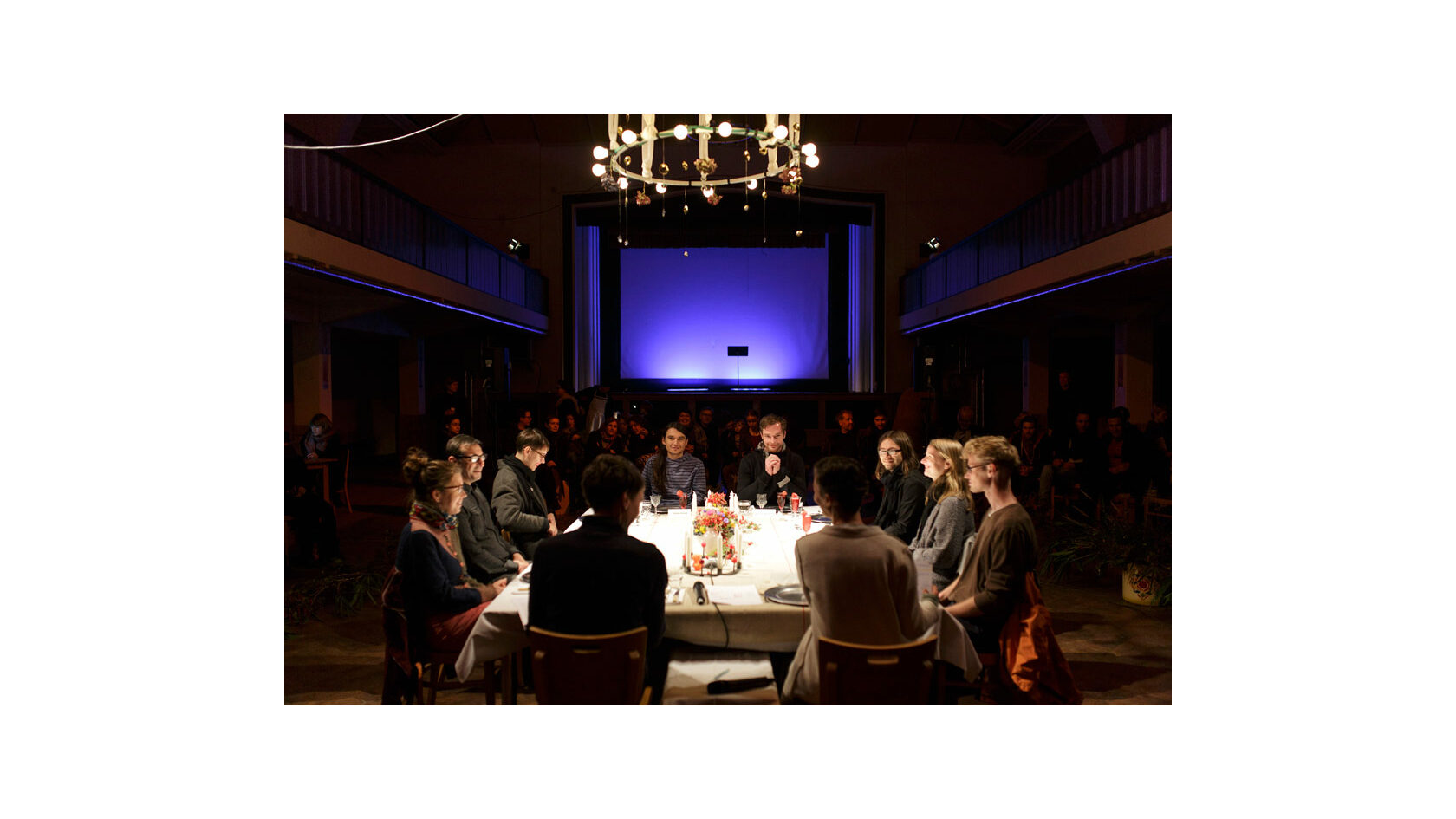 Teilnehmende des Projekts Polylog Dresden sitzen an einem festlich gedeckten Tisch im Ballsaal des Pieschener Zentralwerks, im Hintergrund sitzt das Publikum.