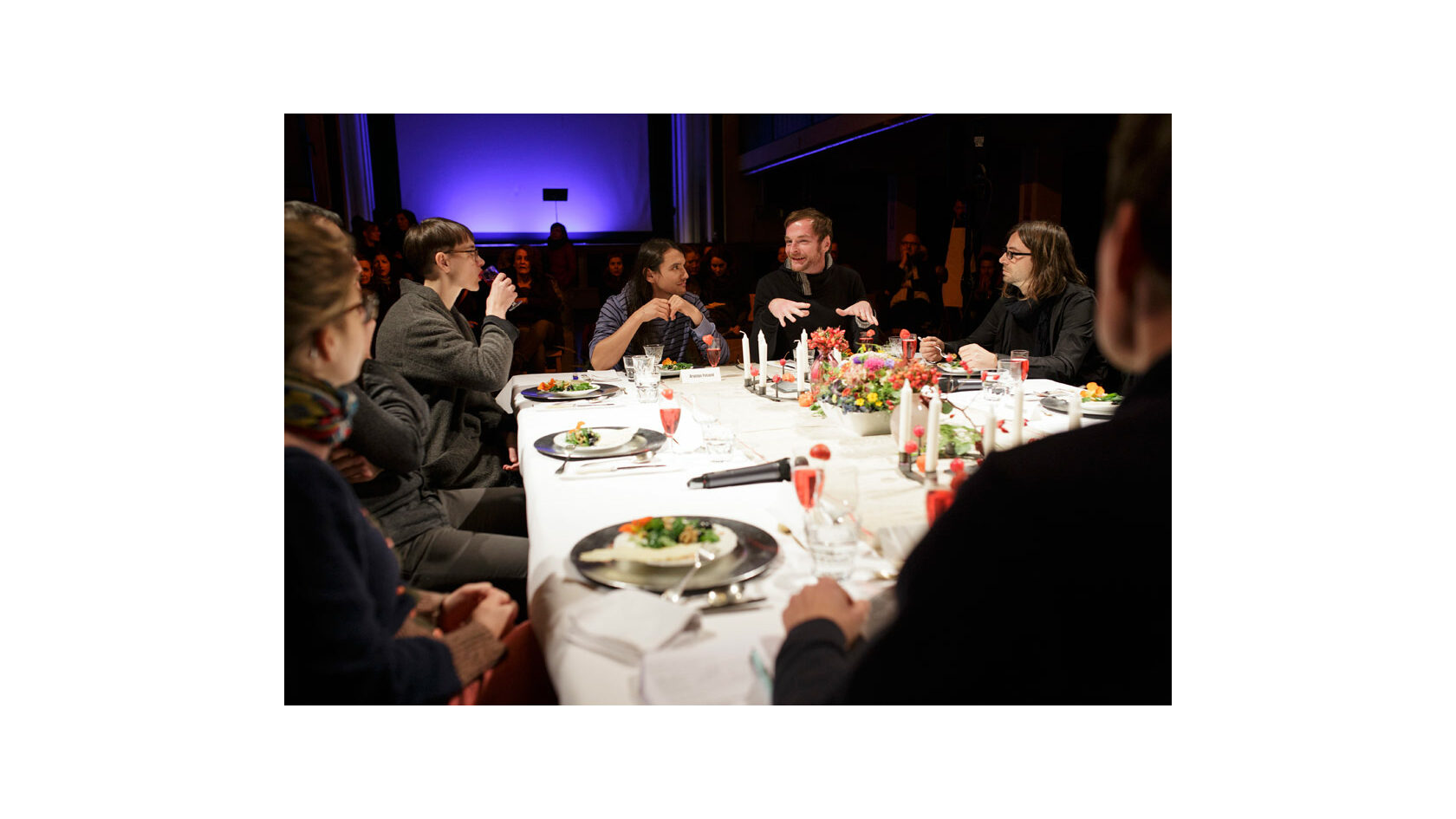 Teilnehmende des Projekts Polylog Dresden tauschen sich während des Abendessens an einem festlich gedeckten Tisch im Ballsaal des Pieschener Zentralwerks aus, im Hintergrund sitzt das Publikum.