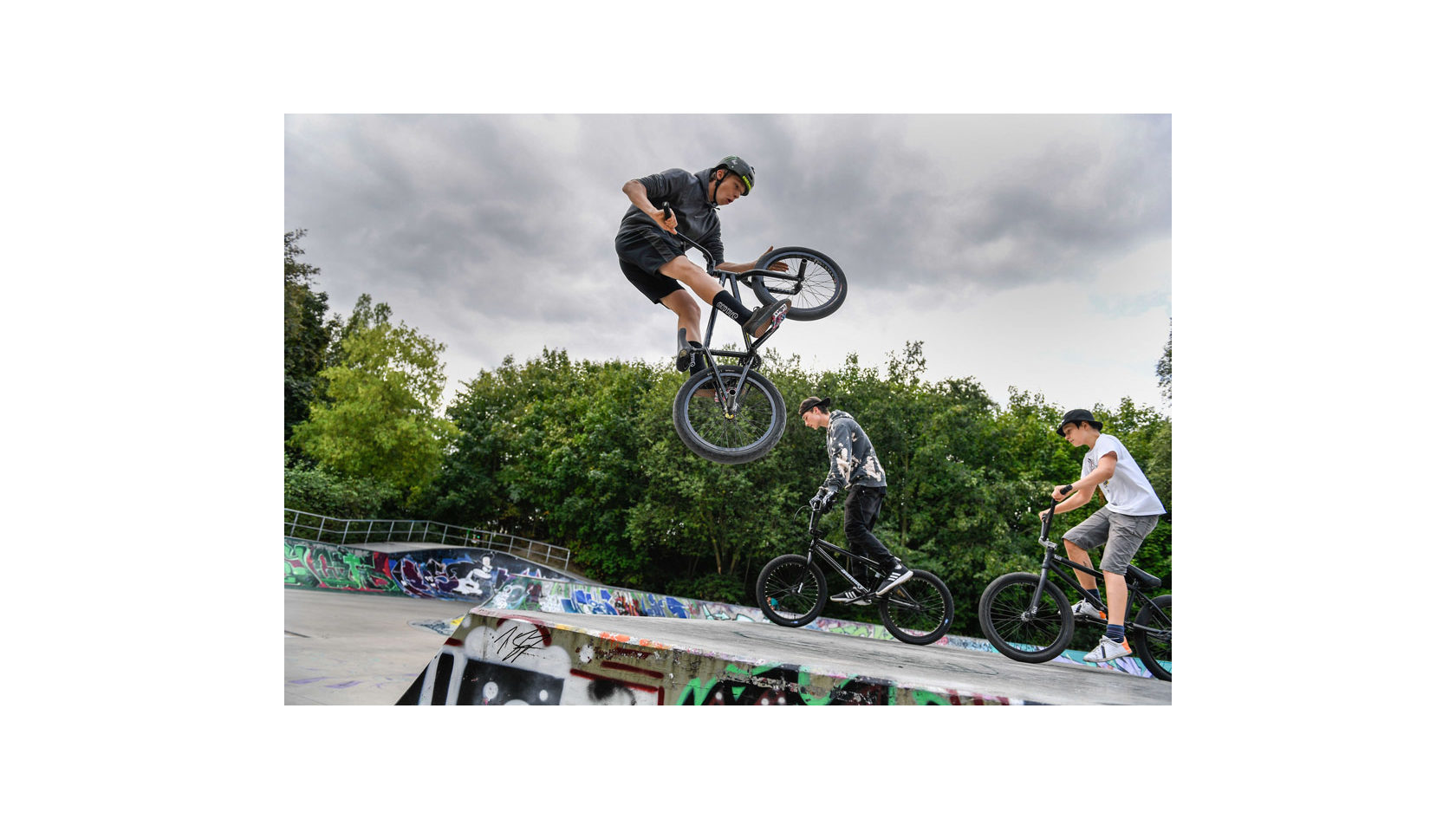 Ein Jugendlicher fliegt mit seinem BMX-Bike auf der Halfpipe durch die Luft, zwei Jugendliche fahren im Hintergrund ihr BMX-Bike.