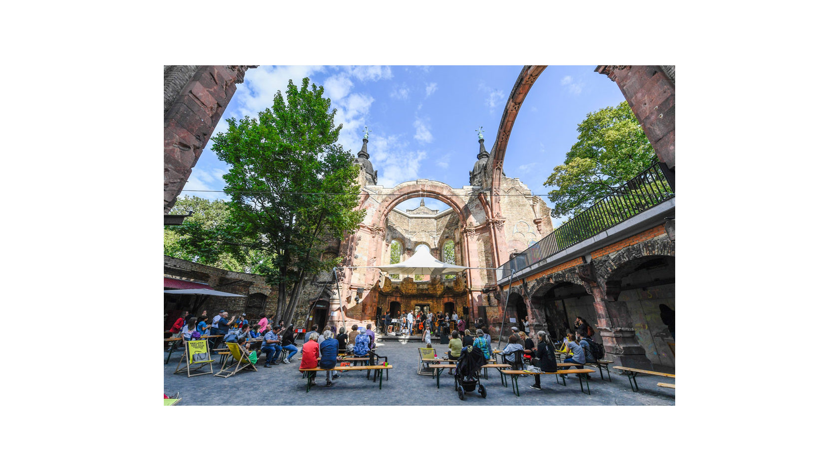 Zahlreiche Menschen sitzen im Innenhof der Trinitatiskirche auf Bänken und beobachten die Aufführung auf der Bühne.