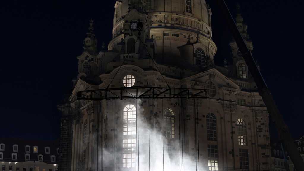 Blick auf die Kunstinstallation Kami auf dem Dresdner Neumarkt vor der Frauenkirche. An einem Kran befestigt schweben fünf menschengroße Figuren, die von Tränengas umhüllt scheinen. Die Installation zeigt Charaktere aus Protestbewegungen der letzten Jahre, die zu Medienikonen wurden.