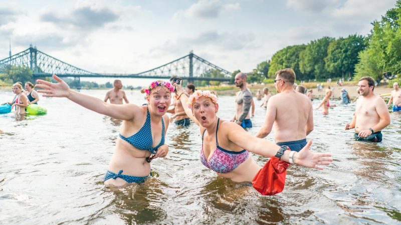 Zwei Elbeschwimmerinnen lachen in die Kamera, während sie im Wasser stehen. Im Hintergrund sind zahlreiche weitere Elbeschwimmer und die Brücke Blaues Wunder zu sehen.