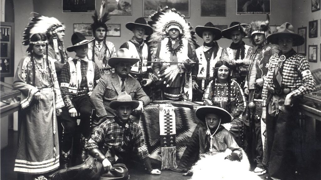 Historisches, schwarz-weißes Gruppenbild von Männern und Frauen, verkleidet als Indianer und Cowboys, versammelt in einem Ausstellungsraum.