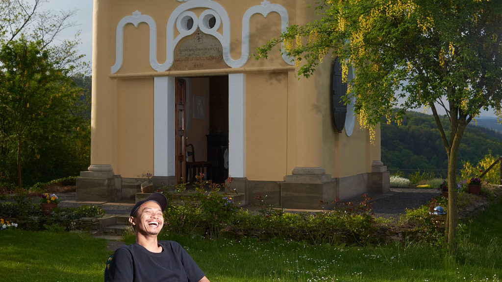 Portrait photo of the Indonesian artist Uji Handoko Eko Saputro who is called Hahan. He sits in a chair in front of the Pavillon in the Saxon village of Maxen.