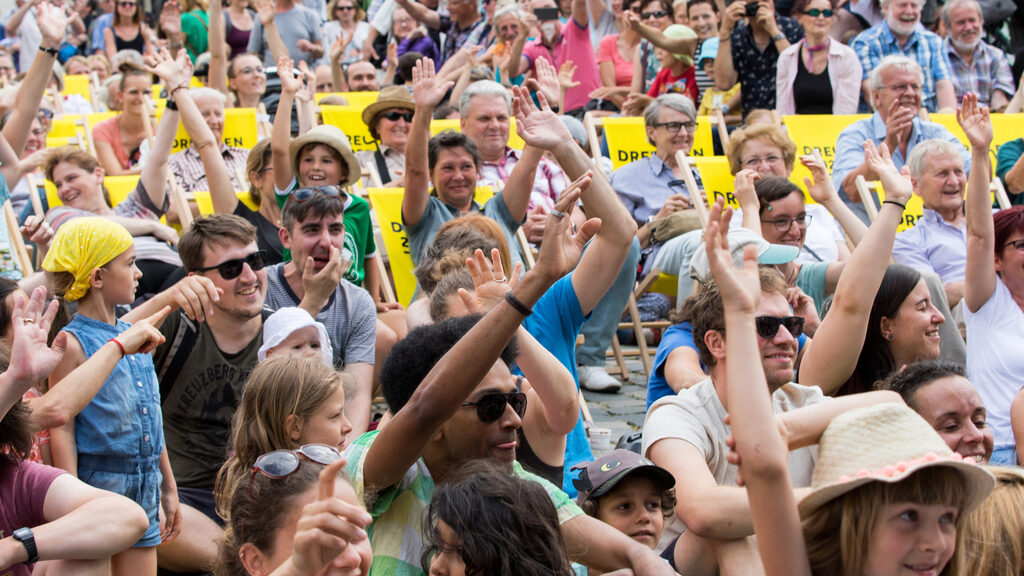 Blick auf das Publikum der Fete de la musique meets Europe. Zahlreiche Menschen heben ihren Arm auf eine Frage hin nach oben.