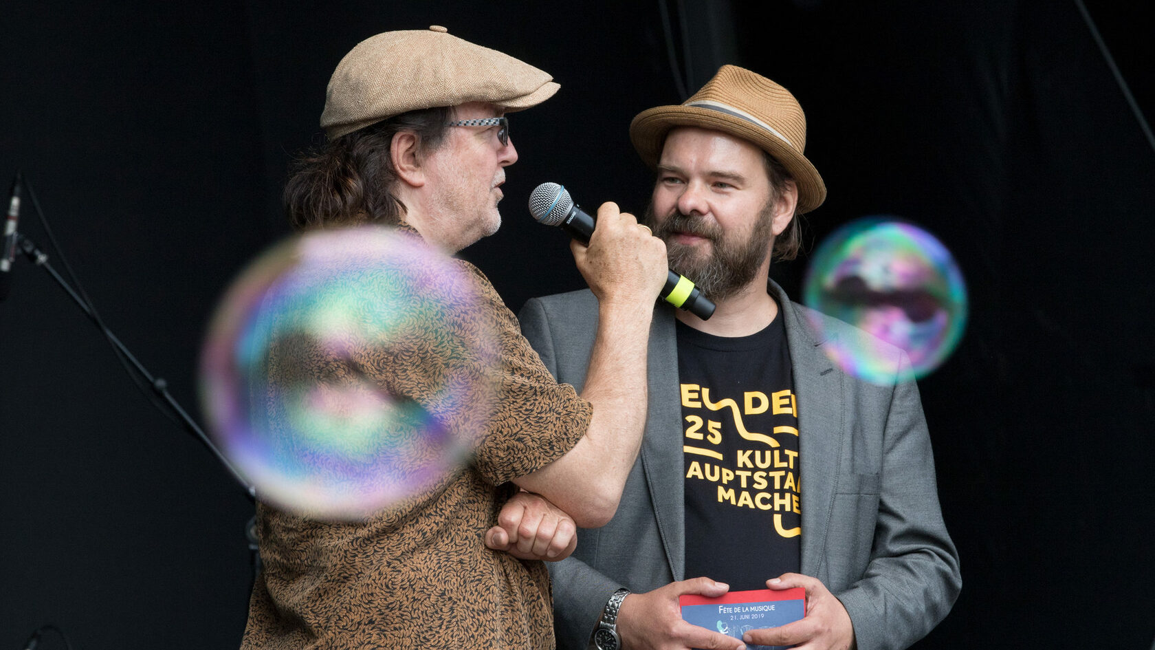 Zwei Künstler unterhalten sich auf der Bühne bei der Fete de la musique meets Europe. Im Vordergrund schweben zwei Seifenblasen.