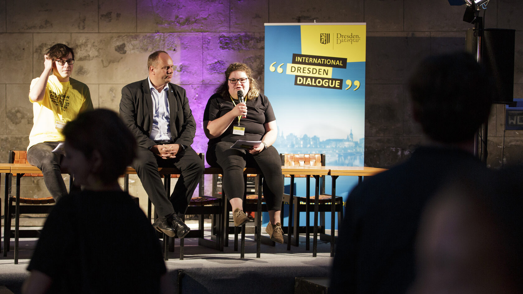 Bei einer Podiumsveranstaltung sitzen Dirk Hilbert, Oberbürgermeister der Landeshauptstadt Dresden, und zwei Jugendliche als Moderatoren auf einem Tisch und diskutieren mit dem Publikum.