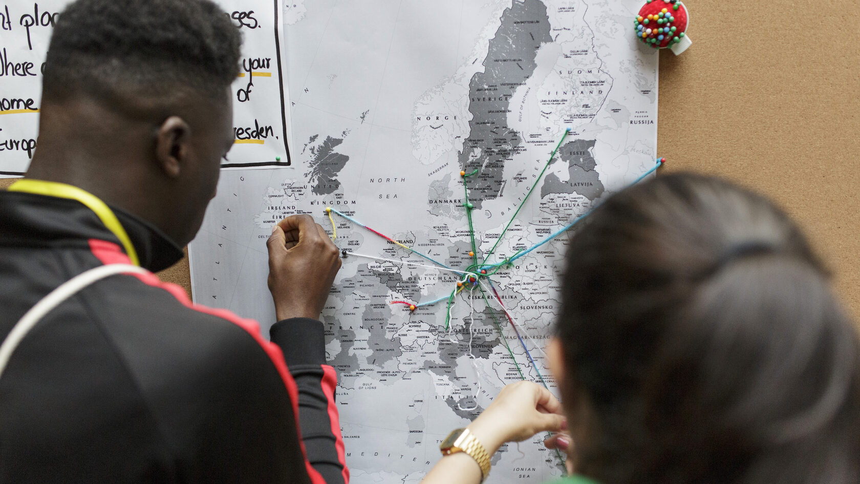 Looking over the shoulder of two young people who are connecting Dresden with other places on a map of Europe using threads of fabric.