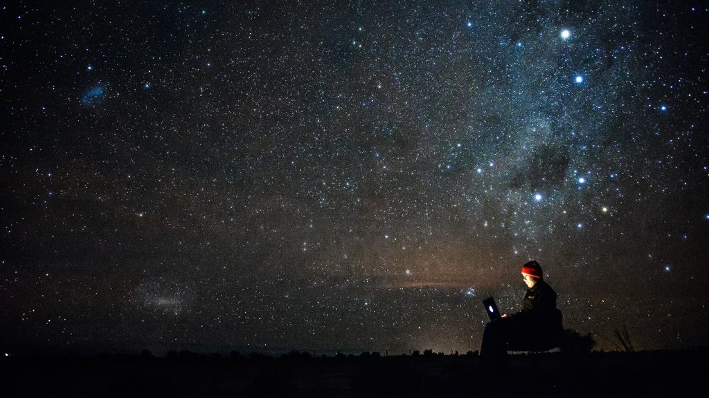 View of a starry sky, in the foreground a man sits in front of a laptop.