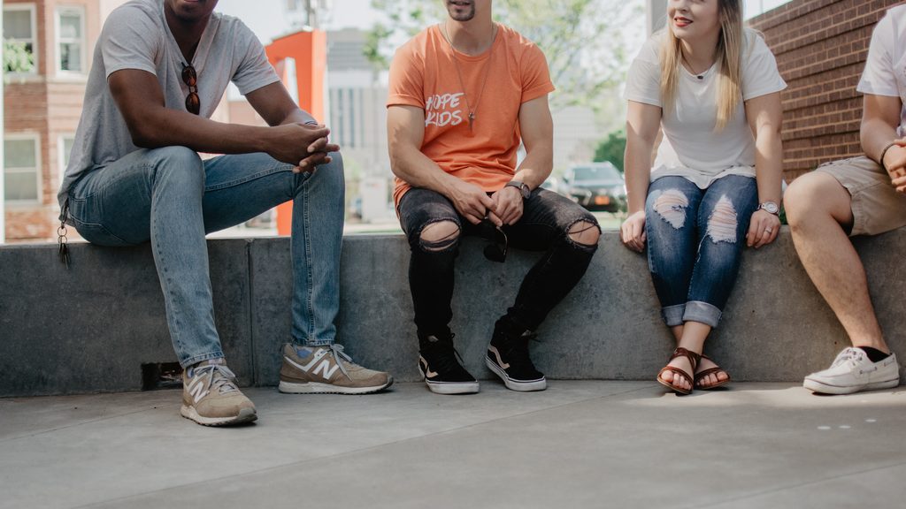 Three men and a woman are sitting on a wall ledge, their faces not visible.