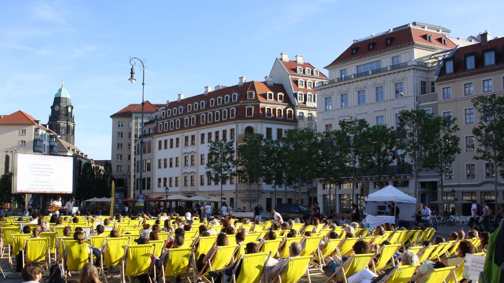 Zahlreiche Menschen sitzen in Liegestühlen auf dem Dresdner Neumarkt und schauen einen Film auf einer mobilen Kinoleinwand.