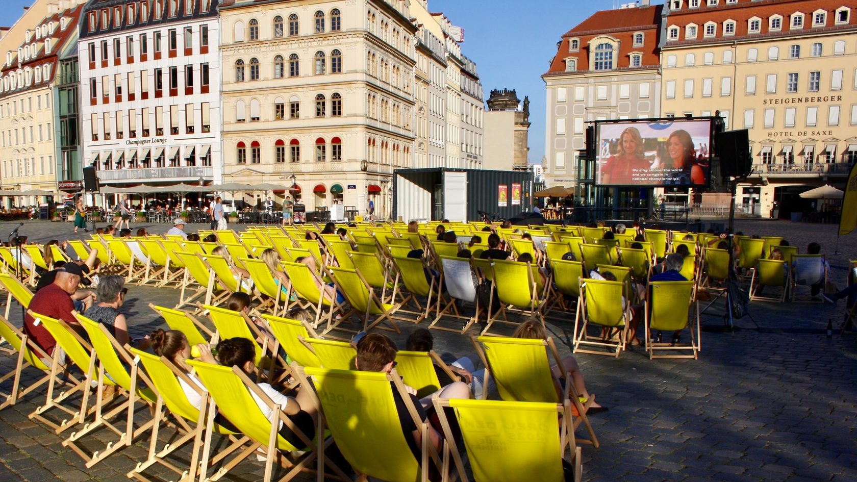 Zahlreiche Menschen sitzen in Liegestühlen auf dem Dresdner Neumarkt und schauen einen Film auf einer mobilen Kinoleinwand.